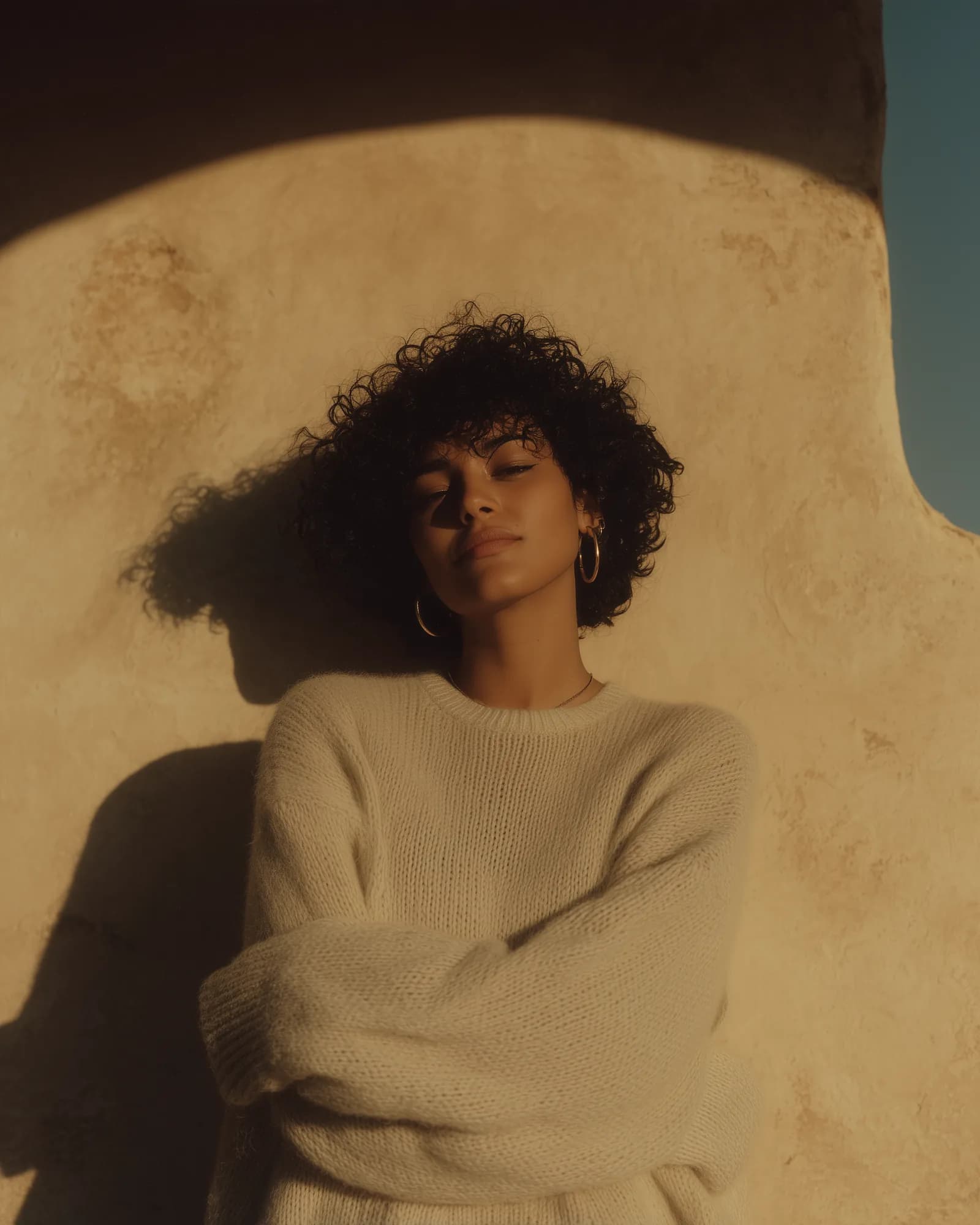 A woman with curly hair, eyes closed, in warm light against a plaster wall